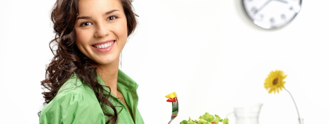 mujer joven sonriendo mientras sostiene en una mano un plato con ensalada y en la otra un tenedor