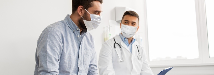 hombre con mascarilla sentado junto a un médico con mascarilla que sostiene carpeta azul en sus manos