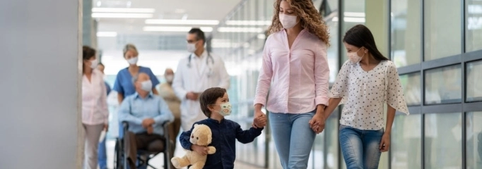 joven madre con blusa rosa, jeans y mascarilla camina por un pasillo de hospital con su hijo y su hija de la mano