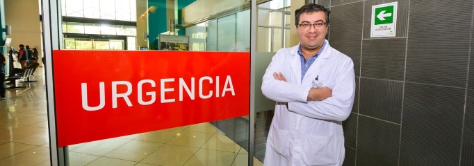 doctor sonriendo delante de una mampara de Servicio de Urgencia, señalética roja 