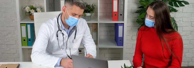 médico con mascarilla y detalantal blanco escribe en su escritorio mientras conversa con paciente mujer joven, de cabello largo y polera roja