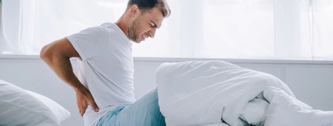 hombre joven sobre una cama con las manos tocando la parte baja de la espalda y rostro que demuestra dolor