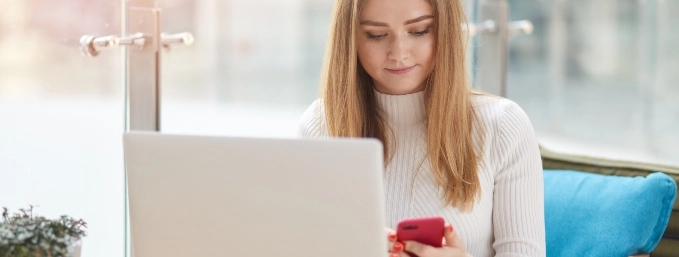 mujer joven mira su celular mientras usa su computador portátil